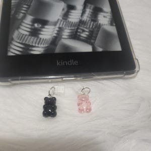 Op de afbeelding: Twee gummy bear bedels, een zwarte en een roze, hangend aan zilveren kettingen. De bedels zijn scherp, terwijl de achtergrond wazig is.