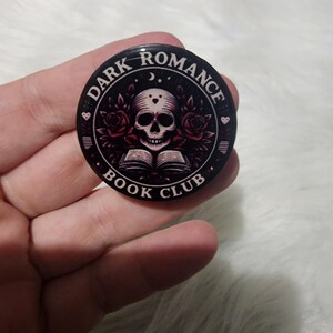 Może przedstawiać: Okrągły, czarny guzik z napisem "DARK ROMANCE BOOK CLUB". Centralny wzór przedstawia czaszkę, róże i otwartą książkę, wszystko w odcieniach czerwieni i bieli.