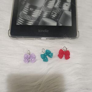 Pode incluir: Três pequenos pingentes de laço em roxo, azul-turquesa e vermelho. Os laços são feitos de plástico e têm um laço prateado para prender a um chaveiro ou bolsa.