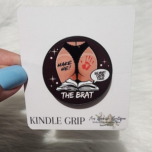 Op de afbeelding: Een ronde Kindle-grip met een afbeelding van een cartoonkont met een handafdruk en de tekst "MAKE ME!" en "DO WHAT YOU'RE TOLD!". De tekst "THE BRAT" staat eronder. De woorden "KINDLE GRIP" staan onderaan.
