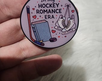 May include: A round, lilac pop socket featuring the text "In my Hockey Romance Era." The design includes a hockey stick, a book, ice skates, and hearts. The pop socket is held in a hand.