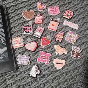 May include: A collection of 18 pink and white bookish charms with various phrases and designs, including "Read More Smut", "Spicy Reader", "Bookish", and "This is my emotional support kindle".