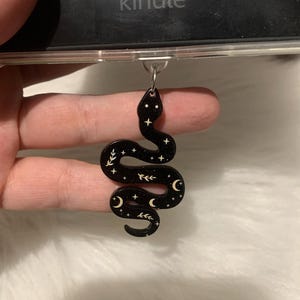 May include: Black snake-shaped charm with a celestial design. The charm features a glossy finish with white stars, crescent moons, and leaf-like accents. It is attached to a silver-colored metal ring, designed to be used as a phone charm.