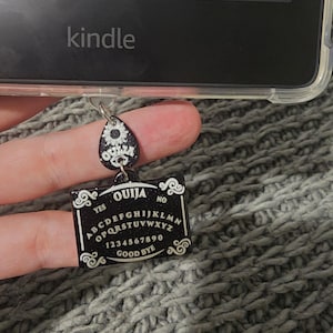 Puede incluir: Un amuleto de tablero de Ouija en blanco y negro con un amuleto de flor en blanco y negro adjunto. Los amuletos cuelgan de una funda de teléfono transparente.