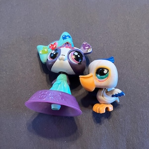 May include: Two colourful Littlest Pet Shop figures. One is a panda with a blue bow and purple accents. The other is a white bird with an orange beak and feet, and blue accents. Both figures have large, expressive eyes.