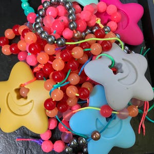 May include: A selection of vibrant beaded necklaces and star-shaped decorations in shades of pink, red, orange, and green. The beads are round and strung, with some necklaces featuring dark grey beads. The star shapes are yellow, pink, blue, and grey.