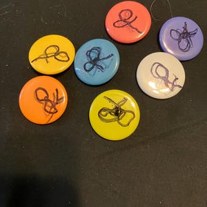 May include: A collection of seven colorful round buttons. Each button is a different color, including yellow, orange, red, blue, white, and purple. Each button features a black, abstract design. The buttons are arranged on a dark surface.