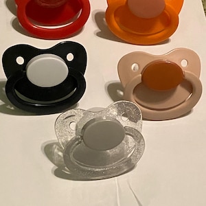 May include: Five baby pacifiers in various colors: red, orange, black, tan, and clear glitter. Each pacifier has a round nipple and a shield with two holes. The pacifiers are arranged on a white surface.