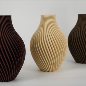 Puede incluir: Tres jarrones con una textura de nervaduras en espiral vertical. El jarrón de la izquierda es marrón oscuro, el central es color crema y el de la derecha es marrón medio. Cada jarrón tiene un cuello estrecho que se ensancha en forma redondeada.