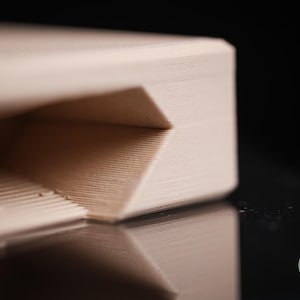 May include: A light beige, 3D-printed object with a smooth, layered texture and a recessed, angular geometric cutout. The surface features fine horizontal lines from the printing process. A small green circular logo reads h3liø design.