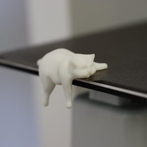 May include: A small, matte white figurine of a cat in a prone, relaxed position. The cat is shaped to hang over the edge of a flat, dark gray surface, with its front paws resting on the top and its hind legs dangling down the side of the edge.