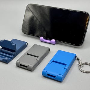 Peut inclure: Un ensemble de supports de téléphone pliables en plastique bleu marine, gris et bleu vif, avec un support violet clair soutenant un smartphone noir. Le support bleu vif possède un anneau porte-clés en métal et tous ont une conception articulée.