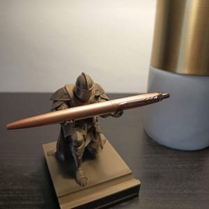 May include: A matte brown knight figurine in a kneeling position holding a metallic rose gold ballpoint pen. The knight is sculpted with detailed armor and a helmet, mounted on a rectangular base, serving as a functional desk pen holder.
