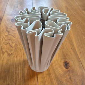 May include: A matte off-white vase featuring a complex, undulating, and pleated structure that creates a series of deep, vertical folds. The top opening displays a rhythmic, wave-like pattern formed by the continuous, curving walls of the vessel.