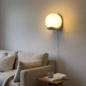 May include: A wall-mounted lamp featuring a spherical, frosted white glass shade that emits a warm, soft glow. The light fixture is supported by a curved, solid white base and includes a thin white power cord extending downward along the wall.