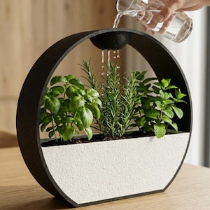 May include: A circular black frame surrounds a white textured planter base containing green basil, rosemary, and mint plants. A small black basin at the top of the frame collects water, which drips down onto the herbs below from a clear glass pitcher.