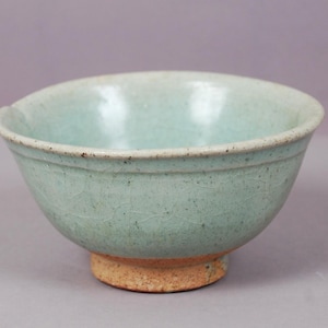 Peut inclure: Un bol en céramique émaillée céladon, de couleur vert clair. Le bol a une forme arrondie avec un bord légèrement évasé et repose sur une petite base non émaillée de couleur beige. La surface présente un motif craquelé.