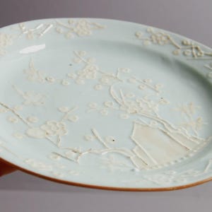 May include: A pale blue ceramic plate with a white floral design. The plate has a raised rim and a slightly scalloped edge.