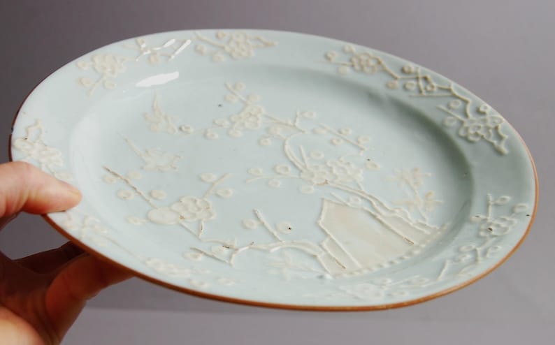 May include: A pale blue ceramic plate with a white floral design. The plate has a raised rim and a slightly scalloped edge.