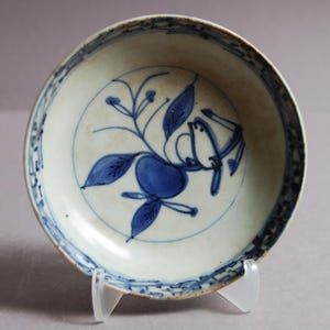 Könnte beinhalten: Kleine, antike weiße Keramikschale mit blauem Blumendesign. Die Schale hat ein zentrales Motiv aus blauen Früchten und Blättern, umgeben von einem blauen Rand. Die Schale wird auf einem transparenten Ständer präsentiert.