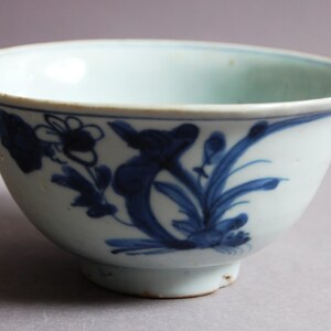 May include: A white ceramic bowl with a light blue interior and a footed base. The exterior is decorated with blue floral and leaf motifs. The bowl has a rounded shape and a slightly flared rim.