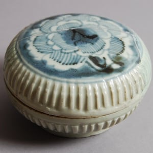 Op de afbeelding: Een kleine, witte keramische pot met deksel, versierd met een blauw en wit bloemenmotief. De pot heeft een geribbeld patroon aan de zijkanten.