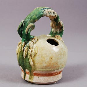 Puede incluir: Un recipiente de cerámica con cuerpo redondo y asa arqueada verde. El recipiente es blanquecino con detalles verdes y marrones. El asa y los elementos decorativos tienen un aspecto texturizado.