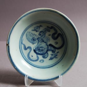 Peut inclure: Petite assiette ronde en céramique antique, intérieur blanc et bord bleu. Le centre présente une illustration bleue détaillée d'un poisson et de plantes aquatiques. L'assiette est soutenue par un support en plastique transparent.