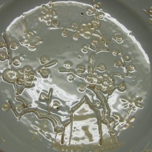 May include: A white porcelain plate with a delicate floral design in a light cream colour. The design features stylised flowers and branches.