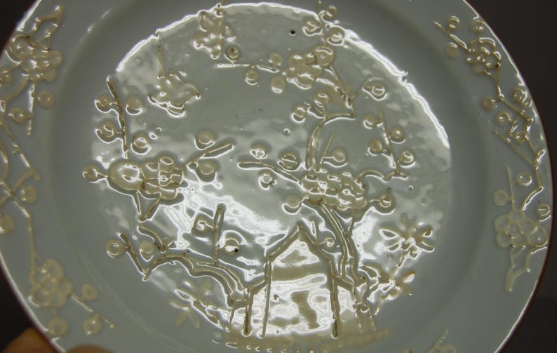 May include: A white porcelain plate with a delicate floral design in a light cream colour. The design features stylised flowers and branches.