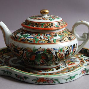 May include: A decorative porcelain teapot with a matching saucer. The teapot and lid are adorned with colorful butterfly and floral patterns. The handle and spout are white, and the lid has a gold finial. The saucer has a scalloped edge.