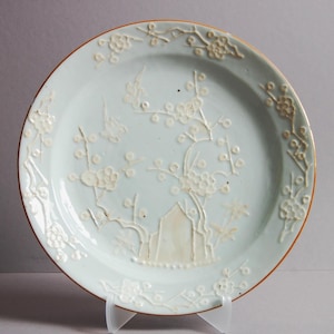 May include: A pale blue ceramic plate with a raised white design of a flowering tree and birds. The plate has a brown rim.