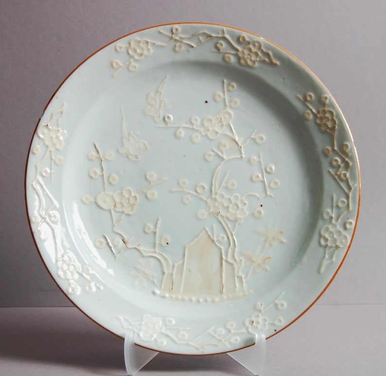 May include: A pale blue ceramic plate with a raised white design of a flowering tree and birds. The plate has a brown rim.