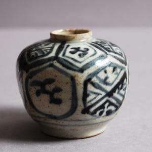 May include: A small, round ceramic vase with a narrow opening. The vase is decorated with a repeating pattern of geometric shapes and stylized motifs in shades of blue and white. The vase is approximately 7.6 cm tall.