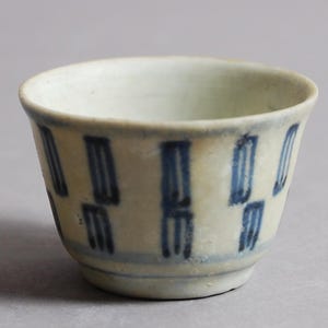 Op de afbeelding: Een klein, wit keramiek kopje met een blauw en wit geometrisch patroon. Het kopje is geglazuurd en heeft een licht afgeronde vorm.