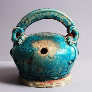 Puede incluir: Un recipiente de cerámica turquesa con asa. El recipiente tiene un cuerpo redondo con un agujero en el centro y un asa arqueada en la parte superior. La cerámica tiene un aspecto envejecido y desgastado con tonos turquesa, beige y rojo.