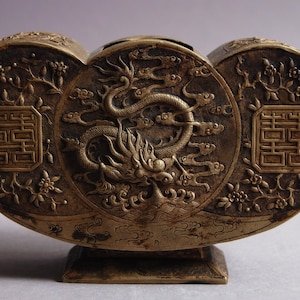 Könnte beinhalten: Ein bronzefarbener, dekorativer Behälter mit Drachen- und Blumendesign. Der Behälter hat eine dreiteilige, herzförmige Oberseite und einen quadratischen Sockel. Aufwendige Details umfassen einen Drachen in der Mitte und chinesische Schriftzeichen an den Seiten.