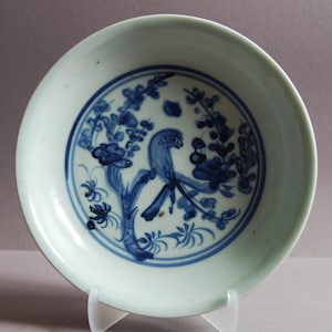 Könnte beinhalten: Kleine, runde, antike Keramikschale mit hellgrüner Außenseite und einem zentralen blau-weißen Design. Das Design zeigt einen Vogel, der auf einem Ast mit Blumen sitzt. Die Schale wird auf einem transparenten Ständer präsentiert.