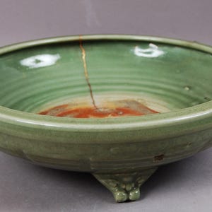 Könnte beinhalten: Eine hellgrüne Keramikschale mit einem breiten, flachen Design. Die Schale weist eine sichtbare Reparatur mit goldfarbenem Material auf, die die japanische Kunst des Kintsugi zeigt. Das Innere hat eine rotbraune Mitte.