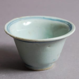 May include: A small, antique porcelain bowl in a pale blue-green colour. The rim features a thin, faded blue band. The bowl has a slightly flared shape and a small, rounded base. The design suggests a vintage or antique piece.