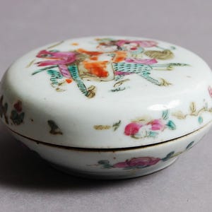May include: A small, white, oval-shaped ceramic trinket box. The lid features a colorful illustration of figures on horseback. Floral and leaf designs adorn the sides. The box is likely vintage or antique.
