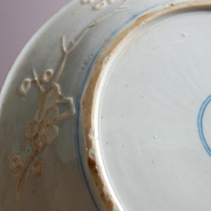 May include: The underside of a white porcelain plate with a blue and white design. The plate has a raised rim and a floral design around the edge.