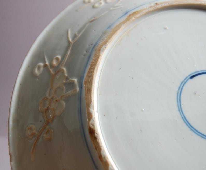 May include: The underside of a white porcelain plate with a blue and white design. The plate has a raised rim and a floral design around the edge.