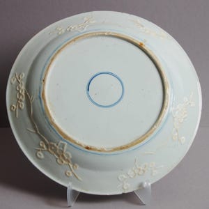 May include: The underside of a white porcelain plate with a blue and white floral design. The plate has a raised rim and a blue circle in the centre.