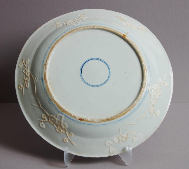 May include: The underside of a white porcelain plate with a blue and white floral design. The plate has a raised rim and a blue circle in the centre.
