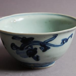 May include: A small, antique ceramic bowl with a pale blue interior and exterior. The bowl has a dark blue, stylised floral design around the outside. The base of the bowl is a darker blue, and the rim is slightly uneven.