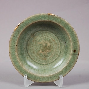 Op de afbeelding: Een celadon-geglazuurde keramische kom met een craquelé-patroon. De kom heeft een afgeronde vorm met een licht verhoogde rand. De kleur is een lichtgroen met gebieden van goudkleurige reparatie. De kom wordt tentoongesteld op een transparante standaard.
