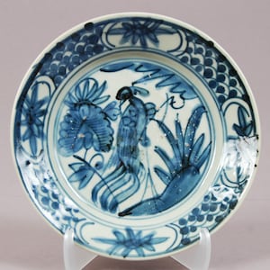 May include: A white ceramic plate with a blue bird and floral design. The plate has a central image of a bird on a branch, surrounded by stylized flowers and foliage. The rim features a repeating pattern of blue dots and floral motifs, typical of traditional pottery.