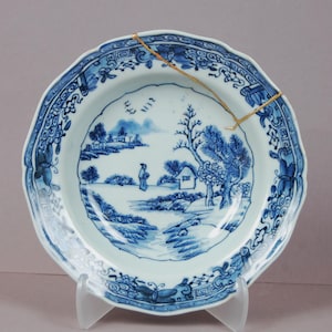 May include: A blue and white porcelain plate with a landscape scene. The plate has a scalloped edge with floral and geometric patterns. The central image depicts a person, trees, and a small house.