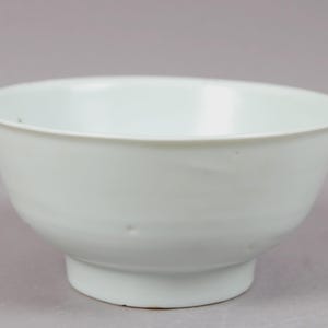 May include: A white ceramic bowl with a rounded shape and a slightly flared rim. The bowl sits on a small, circular base. The smooth surface reflects the light, highlighting its simple design.